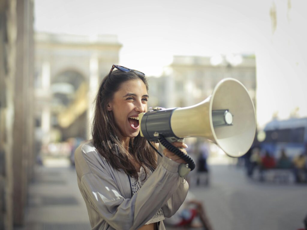 The Power of Words: How What You Speak Shapes Your Reality Cheerful young woman in a casual outfit shouting into a megaphone on a sunny day.